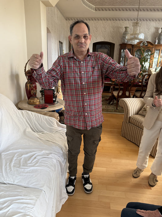 A man shanding with his arms out and giving the thumbs up, he is wearing a red plaid shirt, brown pants, and black and white sneakers. He is in a living room with traditional furniture.